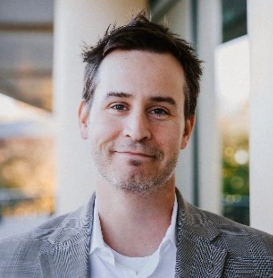 Smiling man in a gray checkered blazer and white shirt, standing outdoors.
